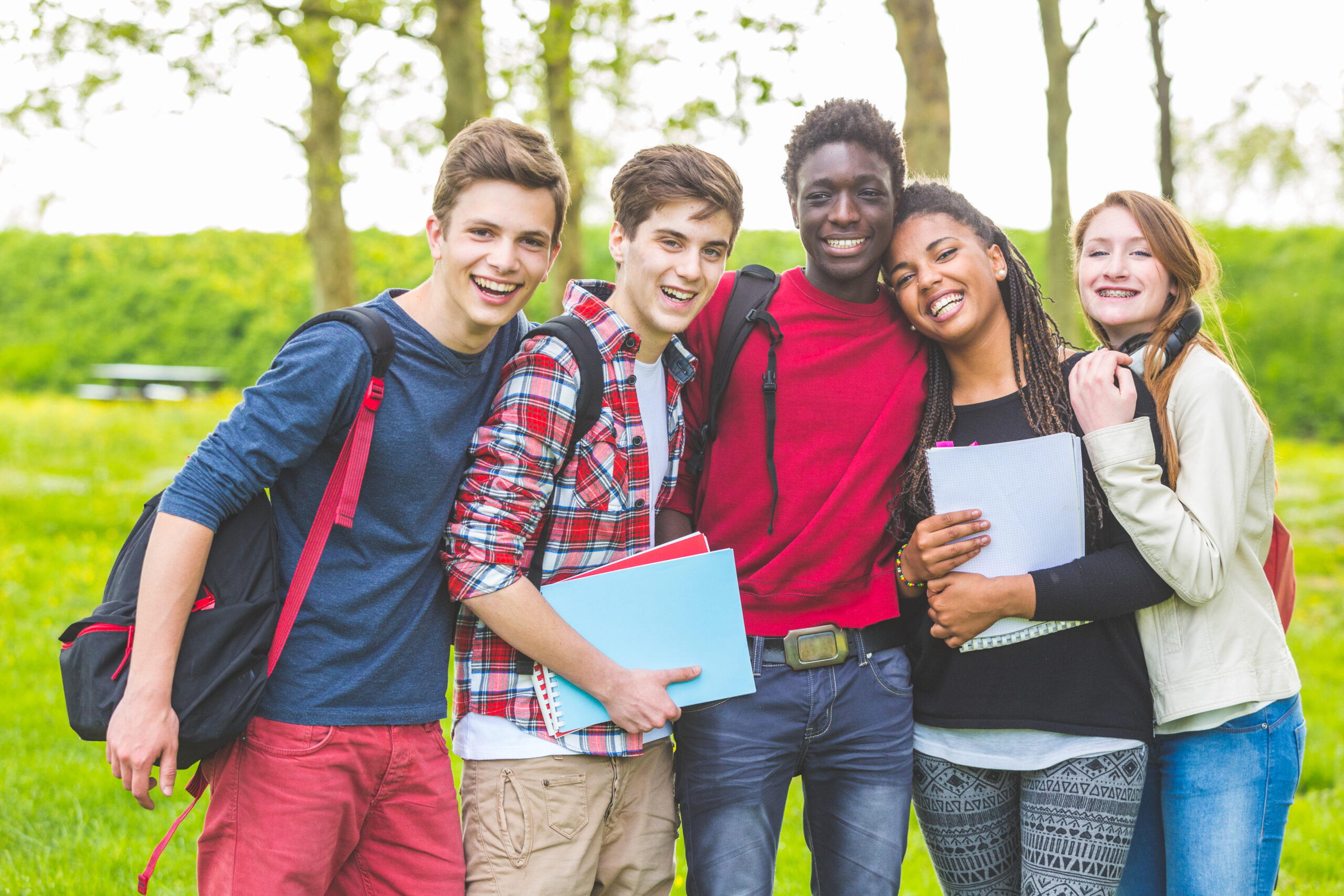 Group of multiethnic teenage students embraced together at park. Two boys and one girl are caucasian, one boy and one girl are black. Friendship, immigration, integration and multicultural concepts.